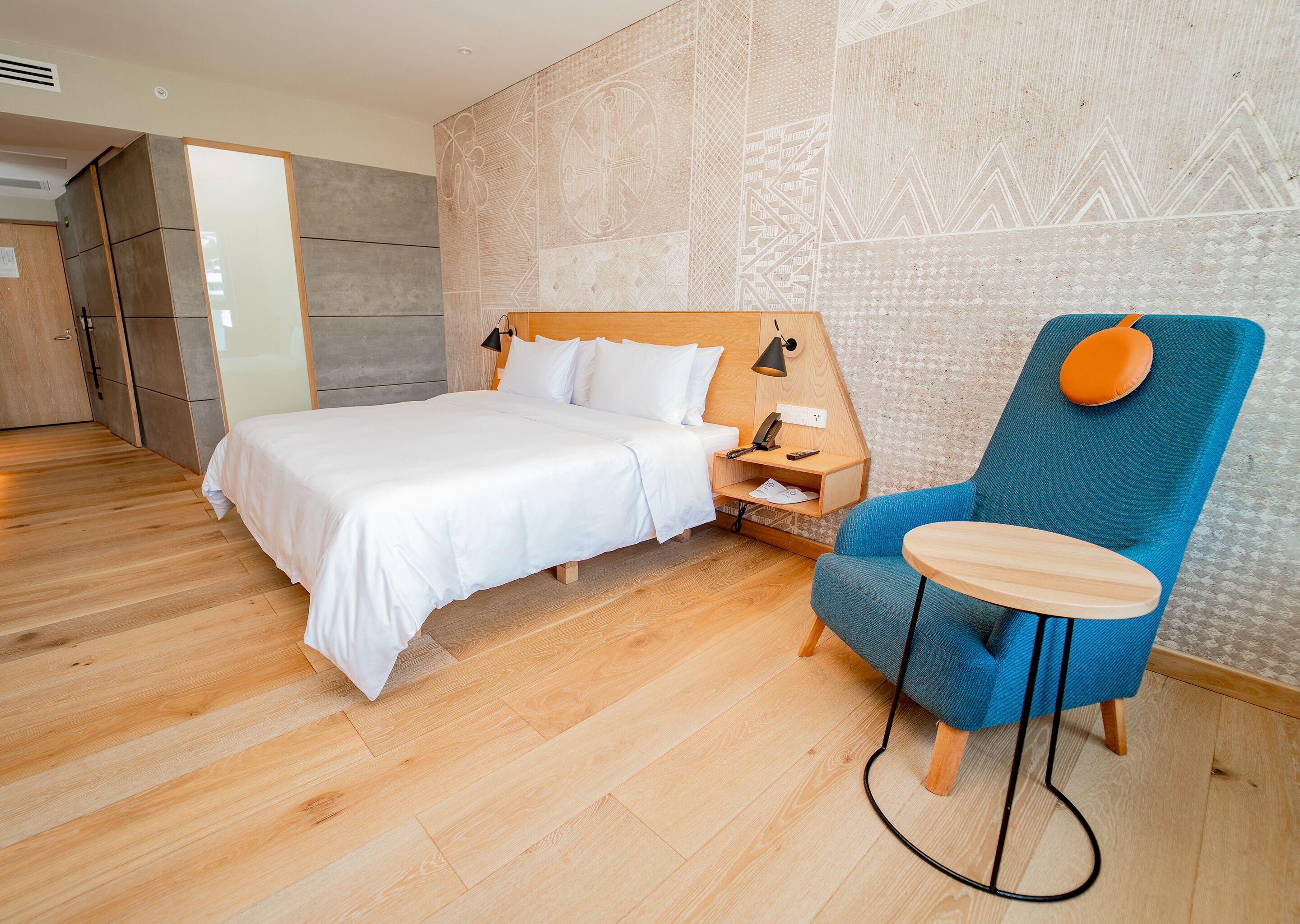 A modern hotel room with a wooden floor, a large bed with white linens, a blue armchair, and a small wooden table.