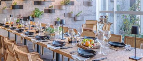 Una larga mesa de comedor dispuesta con sillas, rodeada por una pared con macetas colgantes.