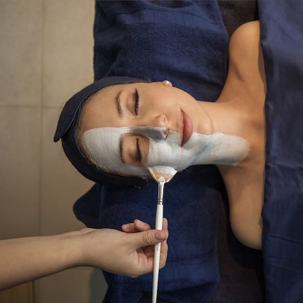 A person lying on a bed with a dark blue blanket, receiving a facial treatment.