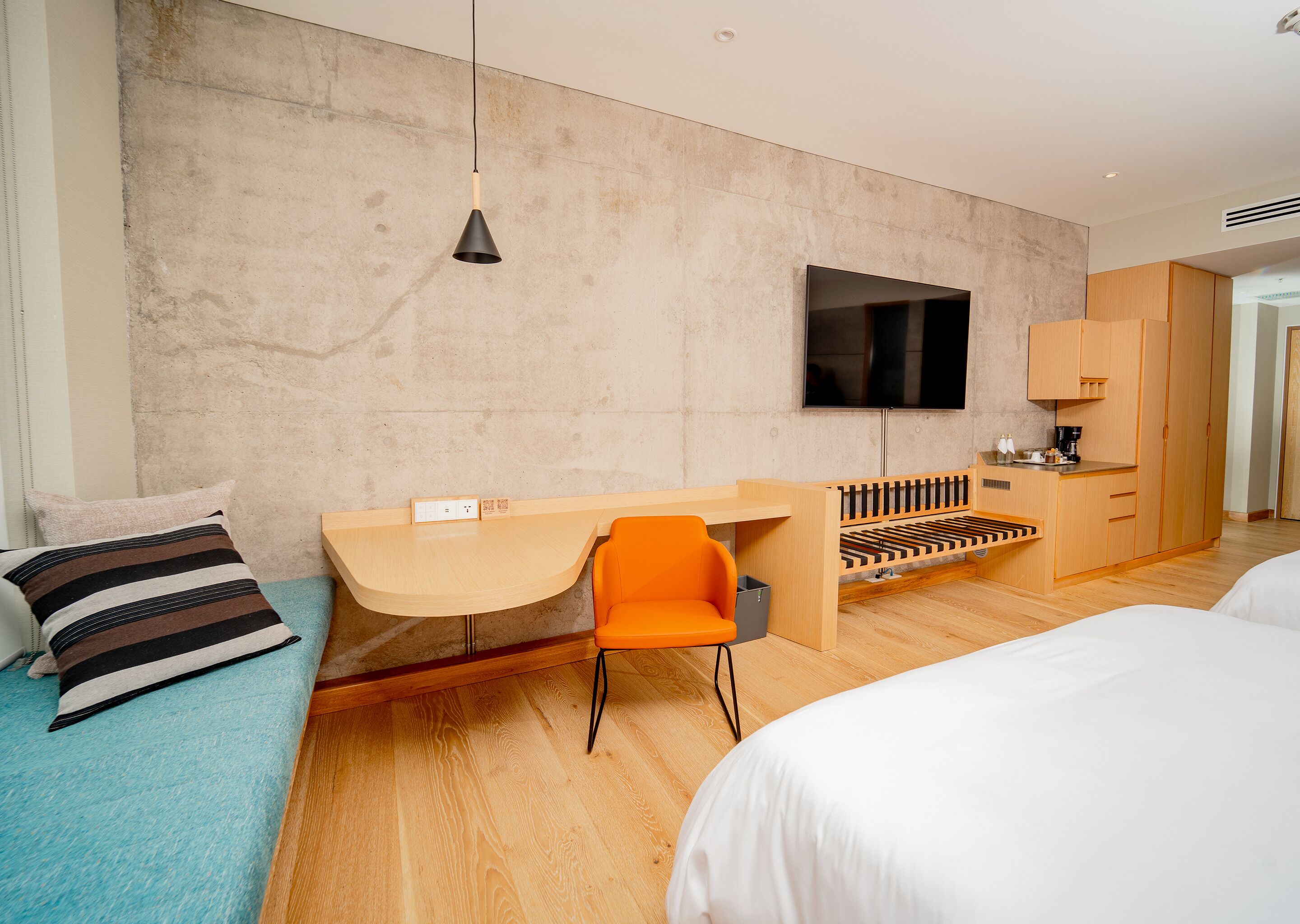 Una habitación de hotel moderna con un escritorio de madera, una silla naranja y una cama con ropa blanca.