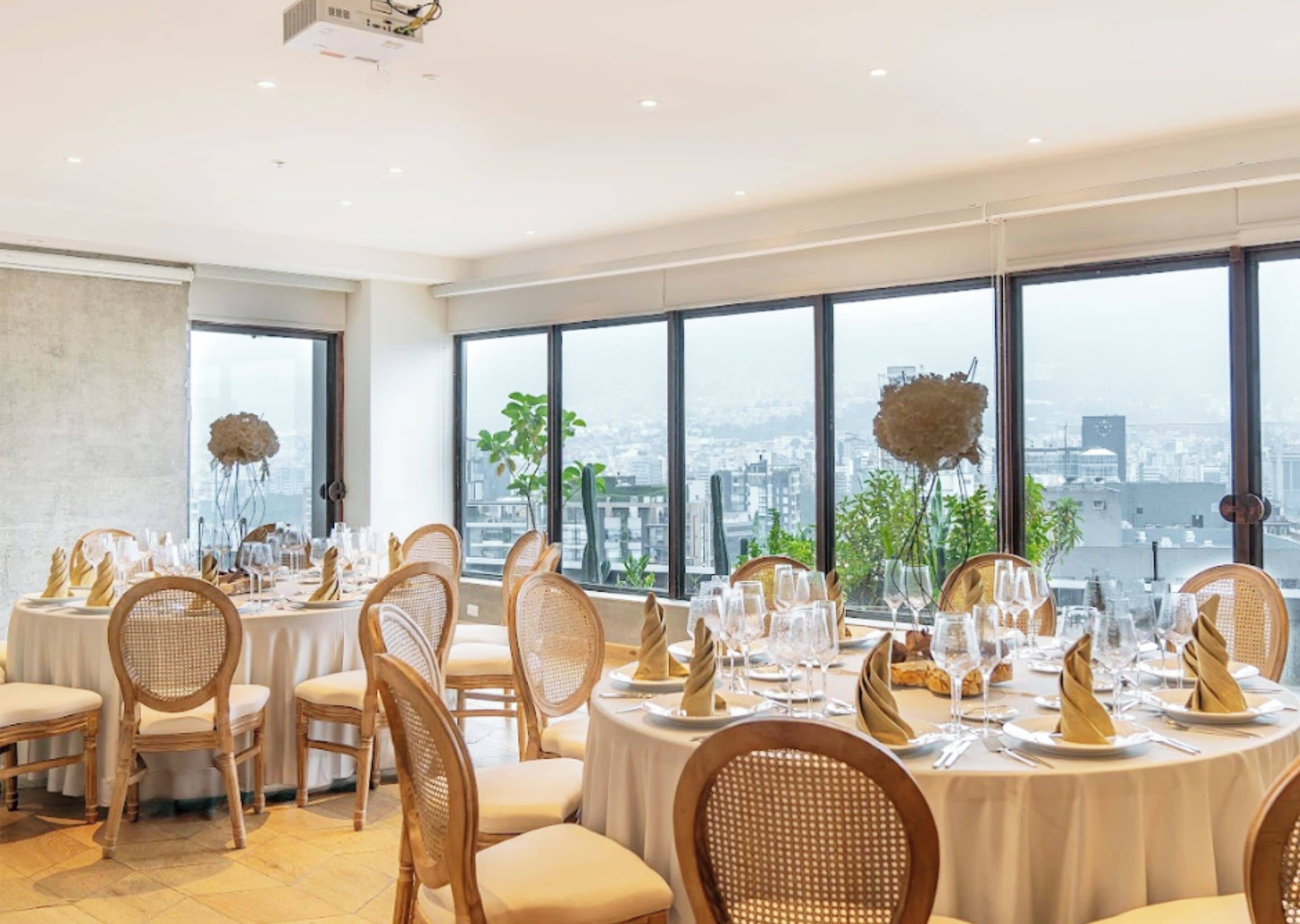 A dining area with round tables set for a meal, wicker chairs, and large windows offering a city view.