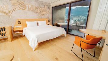 Una habitación de hotel moderna con una cama grande, una mesita, una silla y vistas a una ciudad y montañas a través de una puerta corredera.