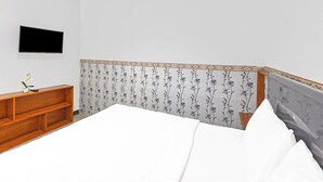 Un dormitorio con cama, televisor montado en la pared, mesita de noche de madera y un cabecero decorativo.
