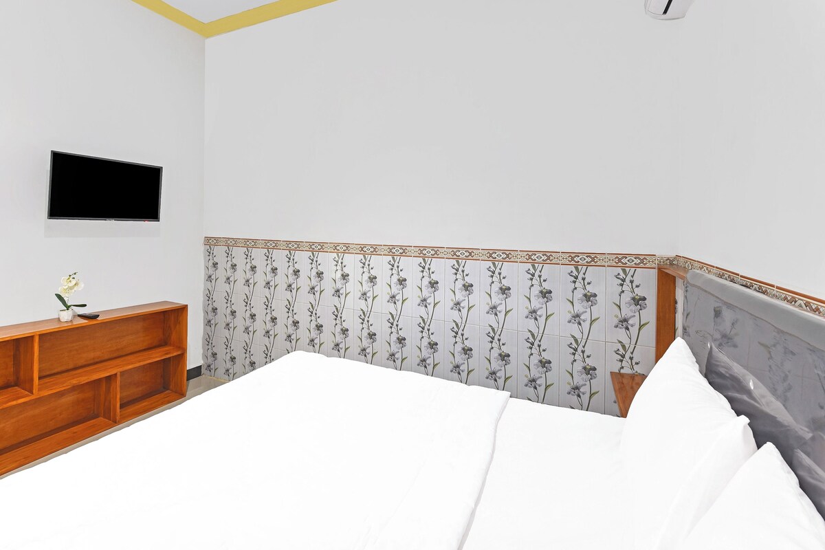 Un dormitorio con cama, televisor montado en la pared, mesita de noche de madera y un cabecero decorativo.