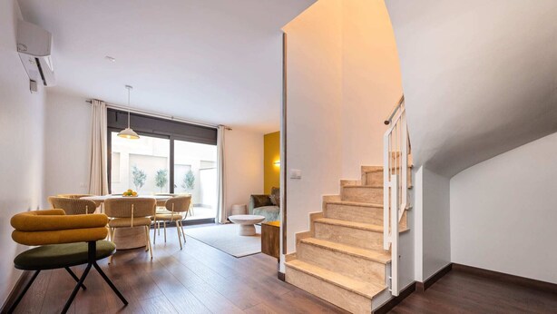 Amplia sala de estar con escalera de madera, comedor con mobiliario de mimbre y un gran ventanal que proporciona luz natural.