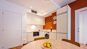 Een compacte keuken met witte kasten, een magnetron, een wasmachine en een eettafel met een schaal fruit.