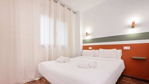 Een hotelkamer met een groot bed, witte lakens, een houten vloer, een gordijn en een muur met een lichtschakelaar en een stopcontact.