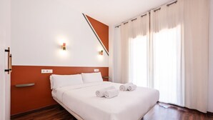 A hotel room with a bed, white bedding, a wooden floor, a wall-mounted light fixture, and a window with curtains. - Sweett | Bailen 104 - Two Bedroom Apartment, Sleeps 4 (Barcelona)