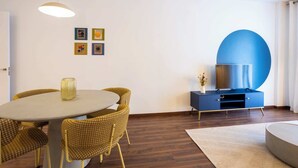 Un salon moderne avec une table à manger ronde, des chaises en osier, un meuble bleu abritant un téléviseur à écran plat et une grande décoration murale bleue.
