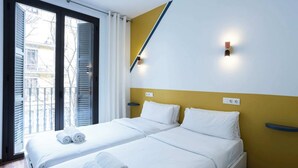 Une chambre d’hôtel avec un lit, deux oreillers et deux serviettes enroulées. On y trouve une fenêtre avec des stores, un luminaire fixé au mur et une prise de courant.