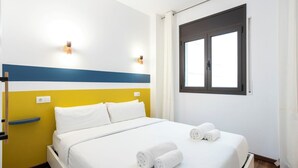 A hotel room with a bed, white bedding, two wall-mounted lights, a window with curtains, and a blue and yellow striped wall design. - Sweett | Bailen 204 - Two Bedroom Apartment, Sleeps 4 (Barcelona)