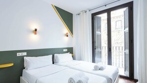 Hotellihuone, jossa on kaksi sänkyä, yksi seinä on maalattu vihreällä korostevärillä, huoneessa on parveke, jossa on sälekaihtimet, ja parvekkeelta on näkymä rakennuksiin.
