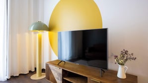 Ein Flachbildfernseher auf einem Holzständer, eine grüne Stehlampe und eine weiße Vase mit Blumen auf einem Tisch.