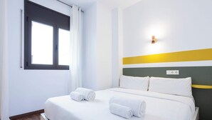 Hotellihuone, jossa on sänky, kaksi rullalle taitettua pyyhettä, ikkuna verhoilla ja seinä, jossa on valokatkaisin.
