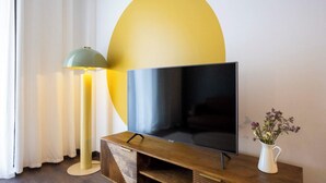 Litteänäyttöinen televisio puisen jalustan päällä, vihreä lattiavalaisin ja valkoinen maljakko, jossa on kukkia, pöydällä.