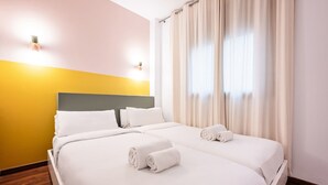 Quarto de hotel com cama de casal, duas almofadas, cabeceira, janela com cortinas e chão de madeira.