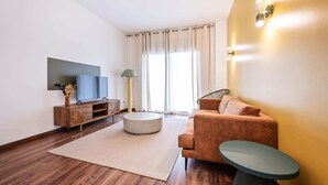 Uma sala de estar moderna com piso de madeira, uma mesa de centro redonda, um sofá, uma TV em um móvel de madeira e um abajur.