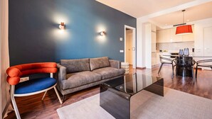 Una sala moderna con un sofá gris, un sillón rojo y azul, una mesa de centro de cristal y una cocina al fondo.
