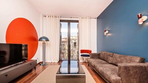Una sala moderna con un televisor de pantalla plana, un sofá gris, una silla rojo y azul, una lámpara de pie y un ventanal con cortinas.