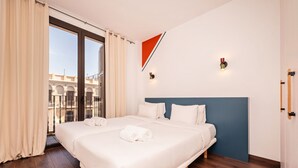 Habitación de hotel con una cama grande, piso de madera, vista a edificios desde un balcón y un adorno triangular rojo en la pared.