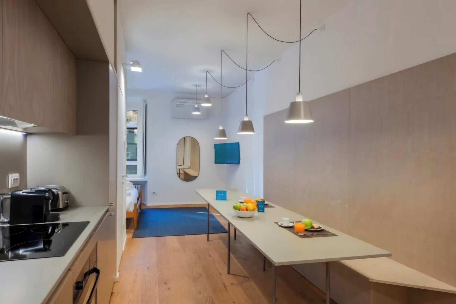 A modern kitchen with a dining area, featuring a long table with fruits and a glass of juice, a toaster, and a stove.