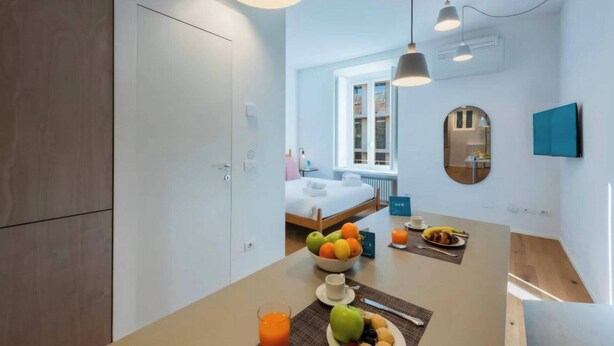 Una camera d'albergo moderna con un letto, un angolo ristoro con frutta e una tazza, e vista sulla città dalla finestra.