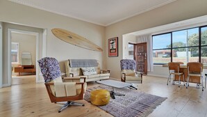 A spacious living room with a large window, a surfboard on the wall, and a cozy seating area with a sofa and armchairs. - A-Wave from it All - 1960's Chic (Warrnambool)