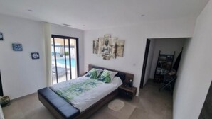Un dormitorio con cama, mesita de noche, una obra de arte en la pared y vista a una piscina a través de un ventanal corredero.