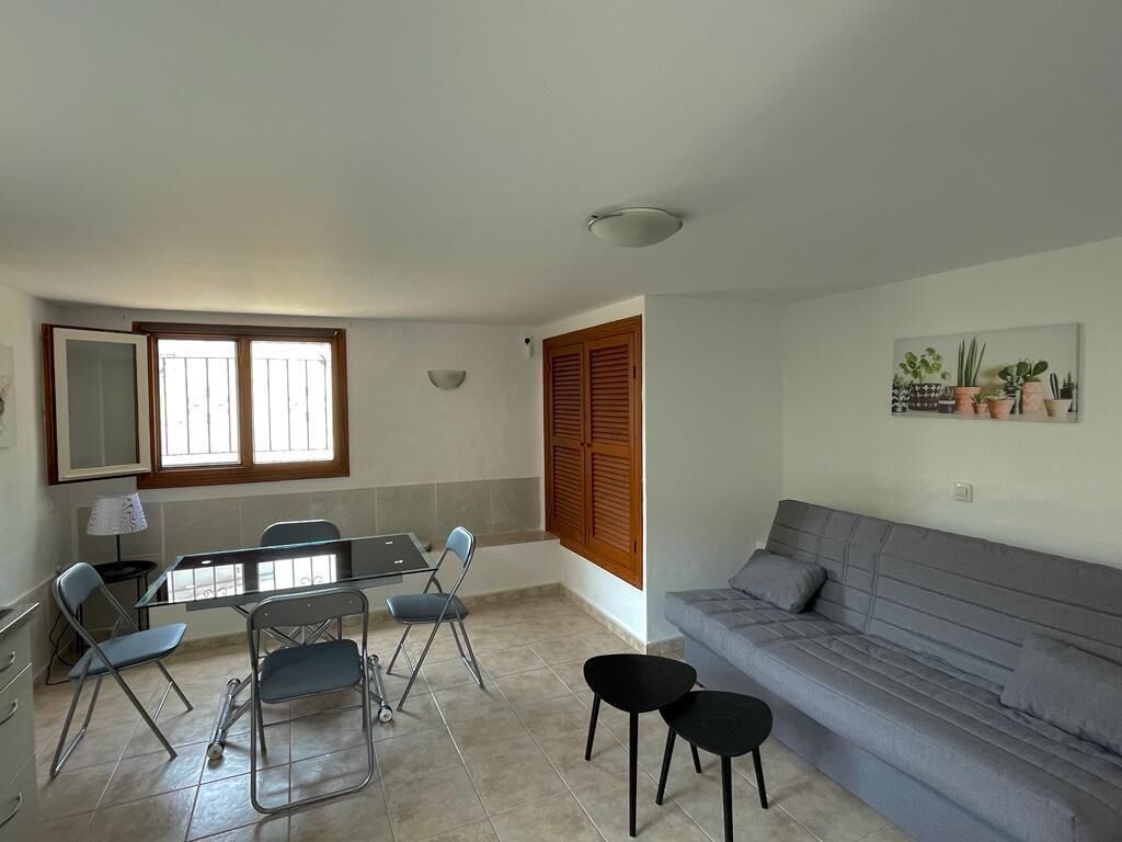 A living room with a grey sofa, a dining table with chairs, and a window with wooden shutters.