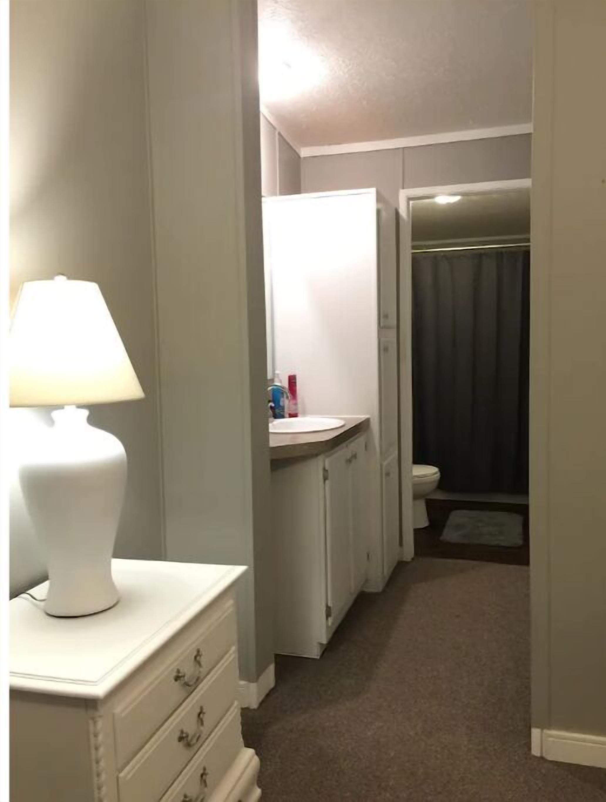 Un couloir étroit avec une commode blanche, une lampe, un lavabo et des toilettes visibles au fond.