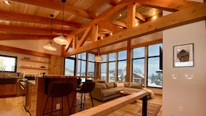 A spacious living area with a wooden ceiling, large windows offering a mountain view, a kitchen with bar stools, and a long wooden bench. - Meridian Lake- Mountain Modern (Crested Butte)