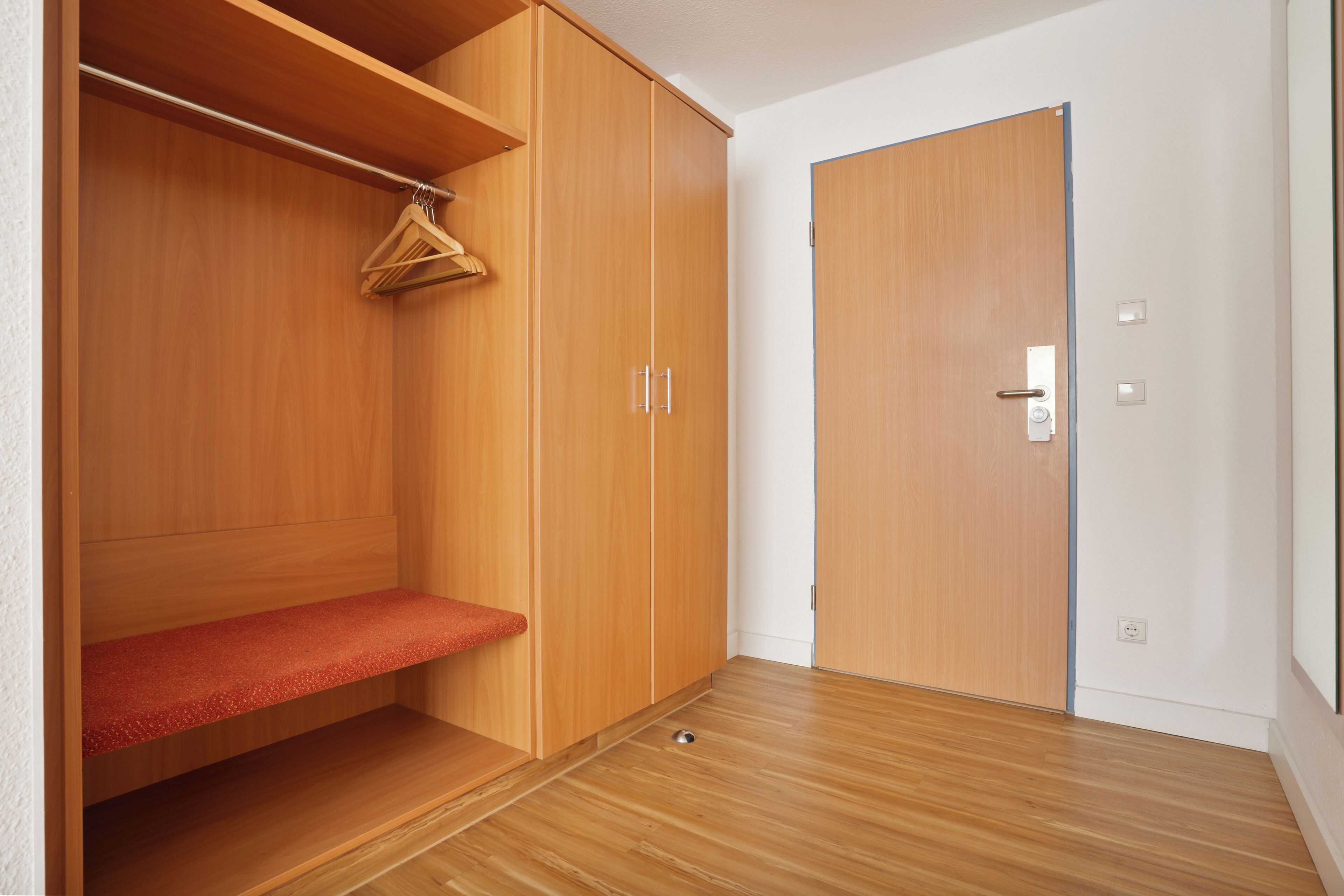 Une pièce avec un plancher en bois, une armoire dotée d’un banc, et une porte avec une poignée.