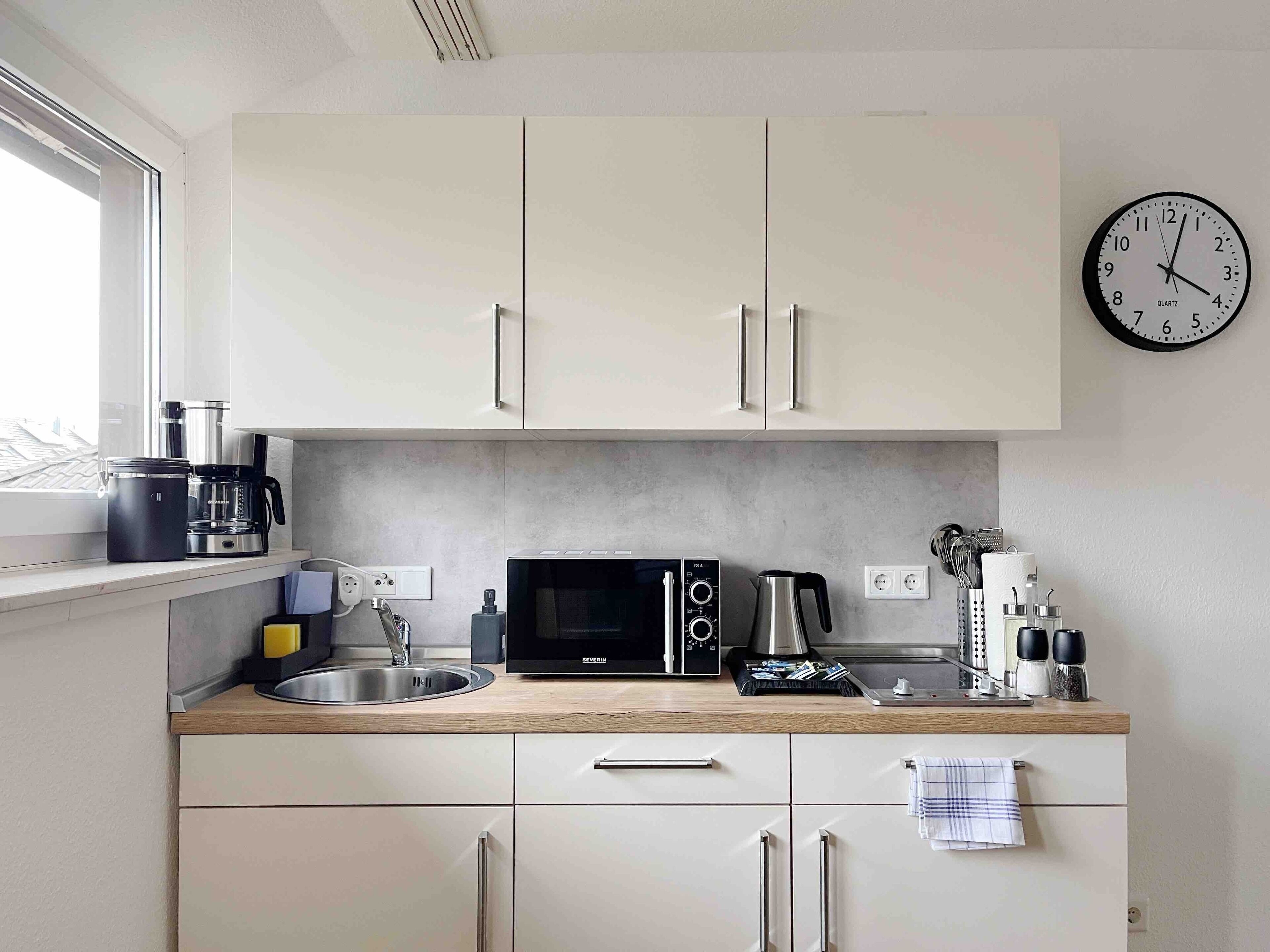 Une cuisine moderne avec des meubles blancs, un four à micro-ondes, une bouilloire et une horloge au mur.