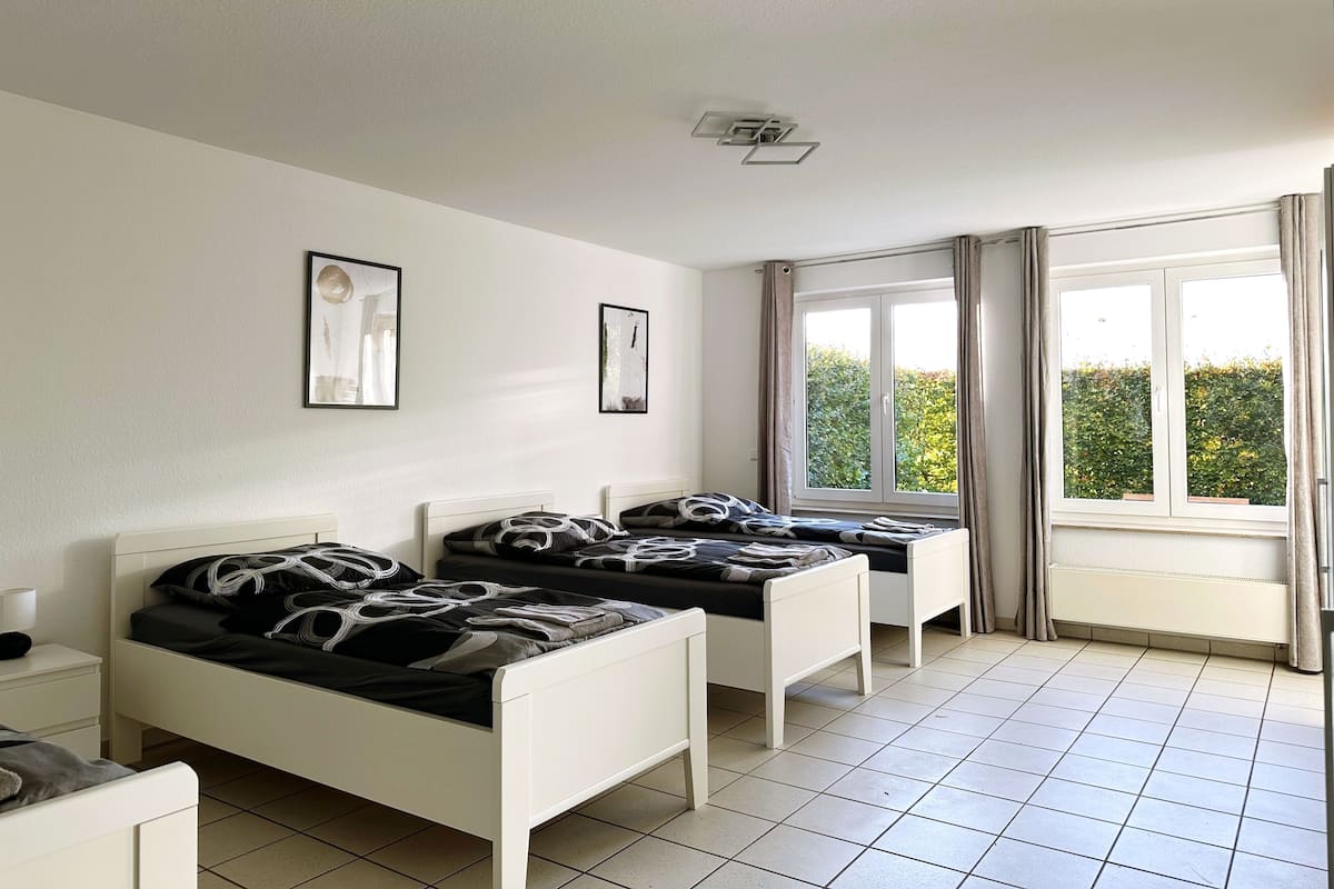 A room with two single beds, a small bedside table, a window with curtains, and a view of greenery outside.