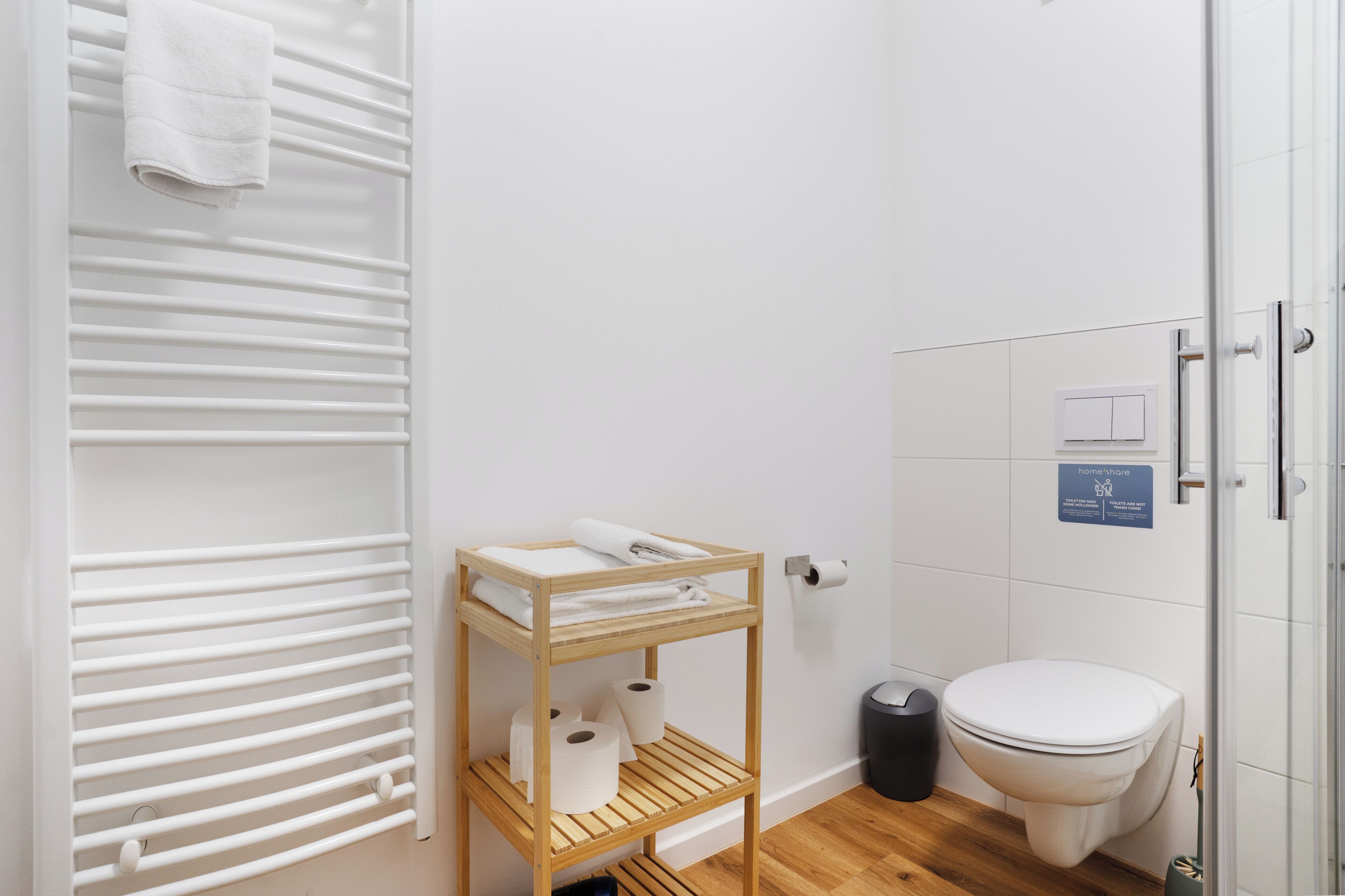 Un baño con un estante de madera que sostiene toallas y papel higiénico, un inodoro blanco y una toalla blanca colgada en un perchero.