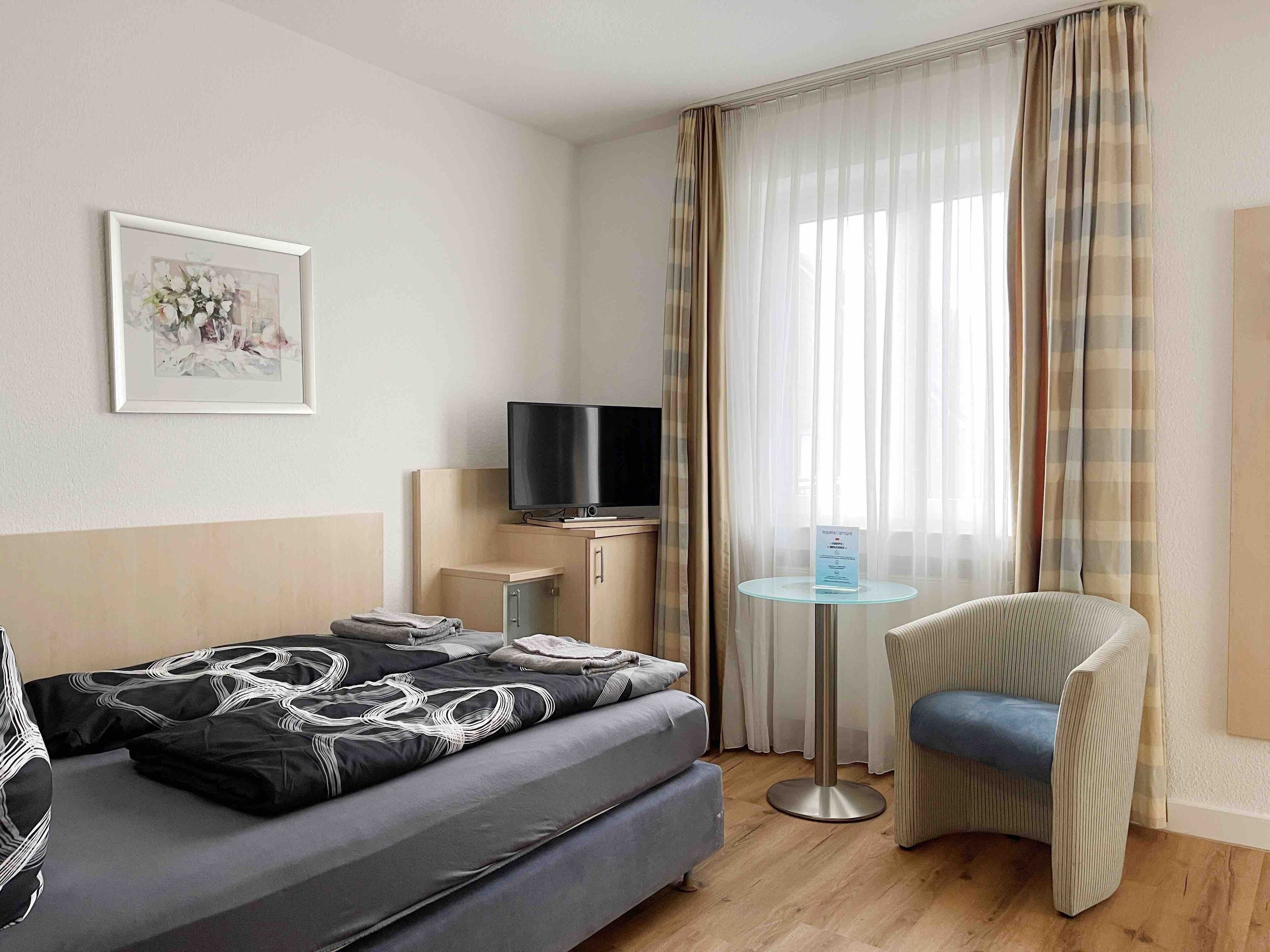 Habitación de hotel con cama, televisión, mesita, una silla y ventana con cortinas.