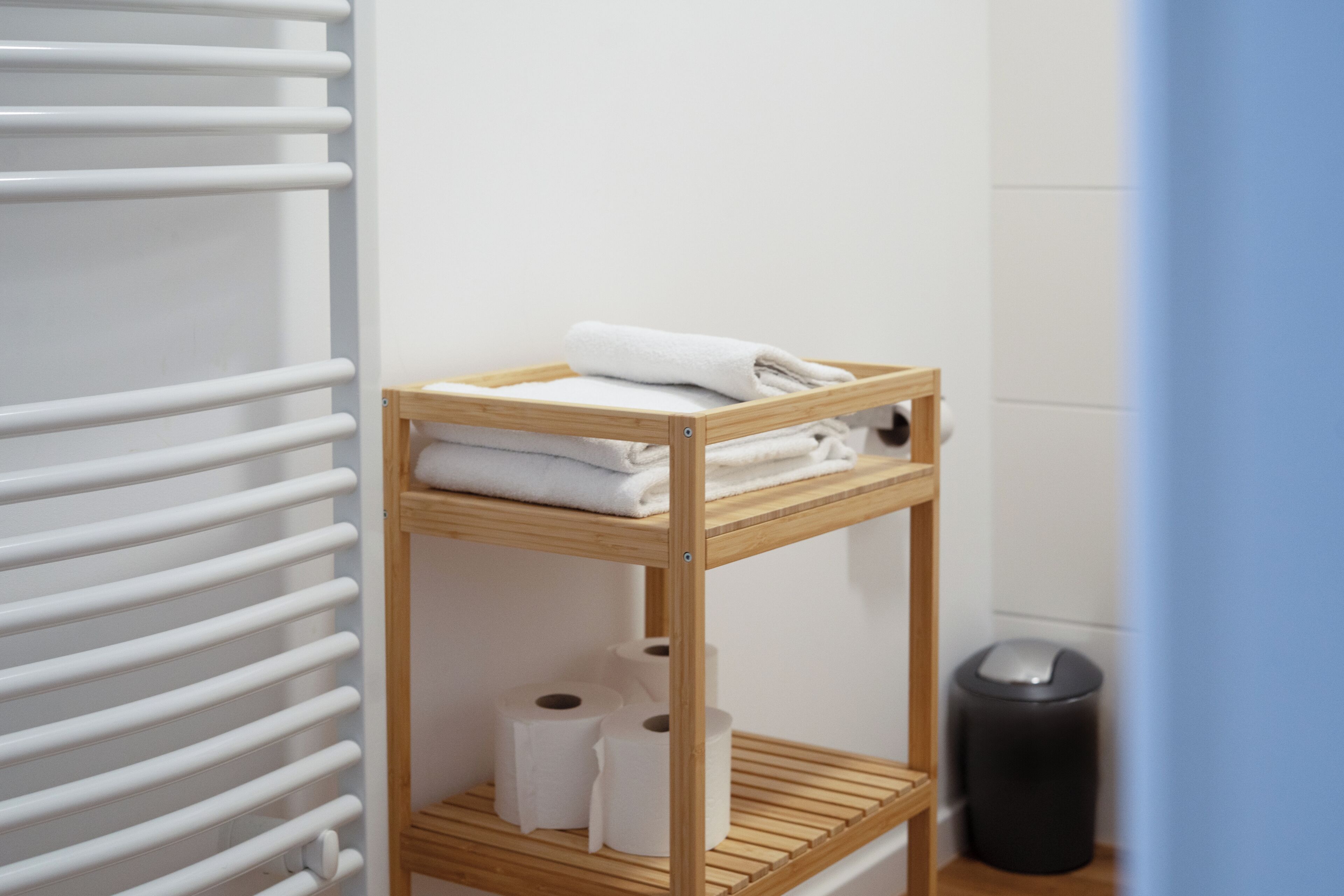 Ein Badezimmer mit einem Holzregal, das Handtücher und Toilettenpapier beherbergt, einem weißen Heizkörper und einer schwarzen Mülltonne.