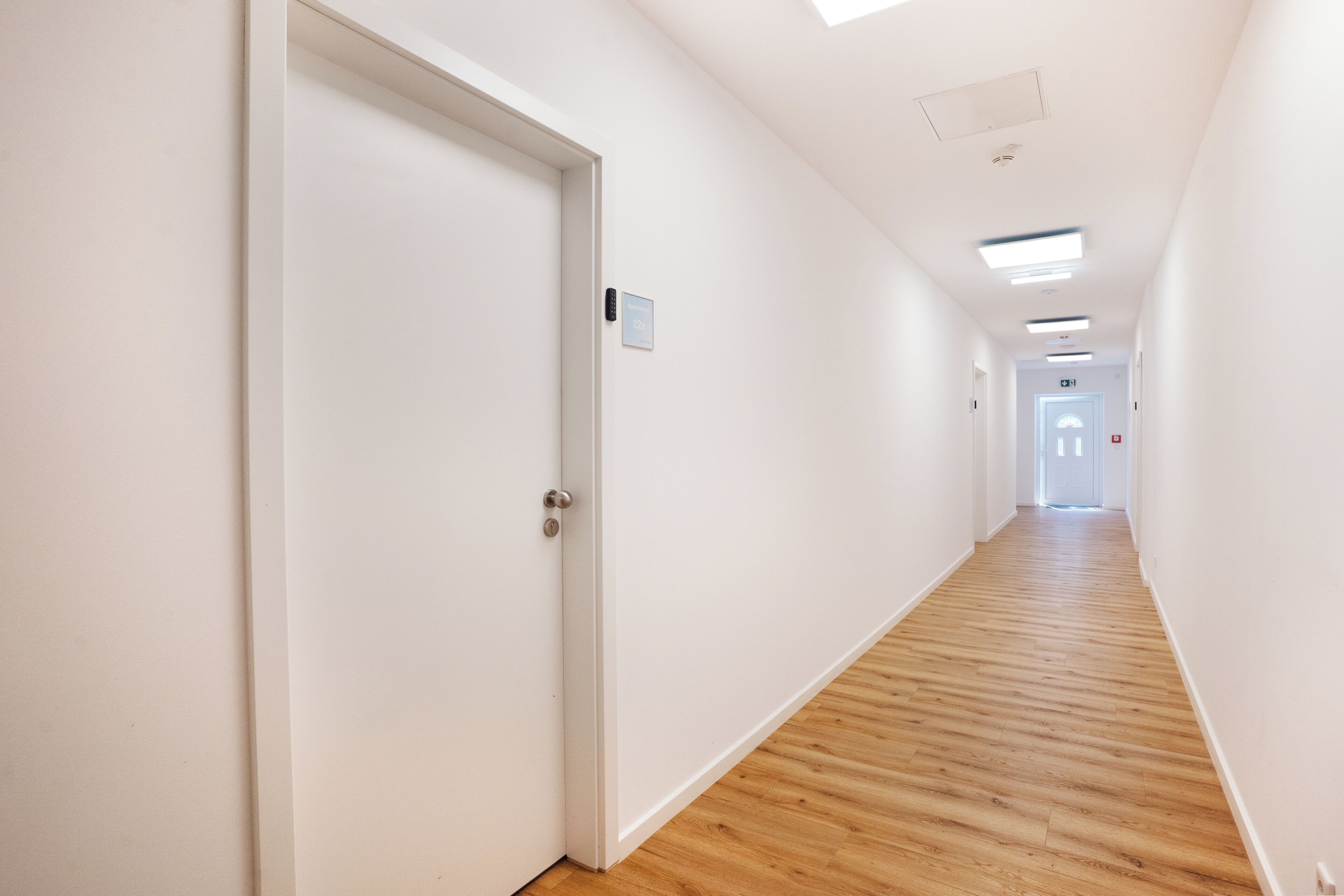 A corridor with white walls, wooden flooring, and a door with a sign.