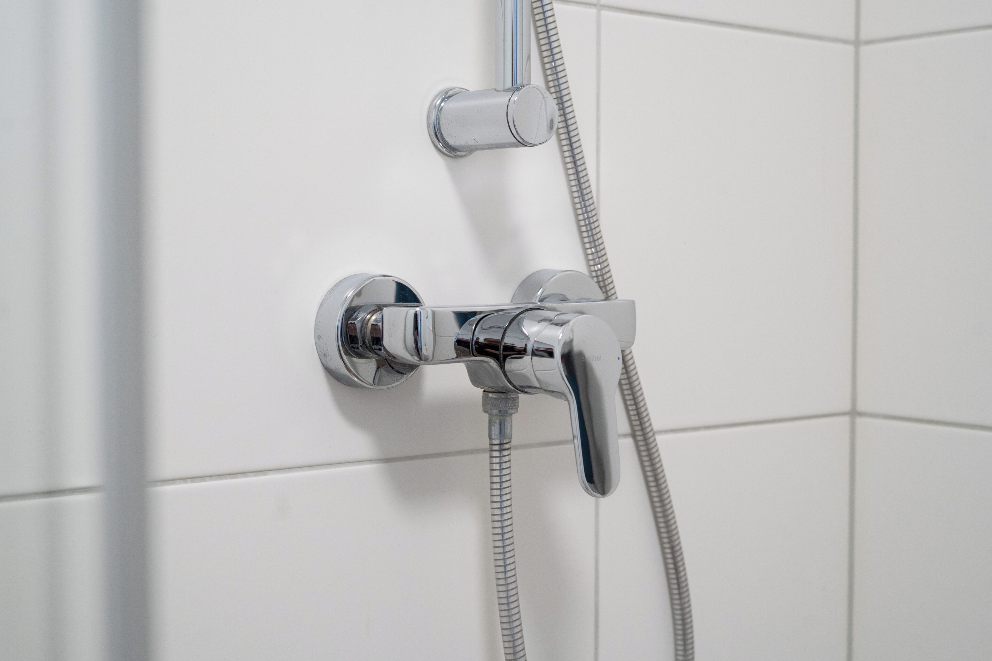 Une pomme de douche chromée, dotée d’un flexible, est fixée à un mur recouvert de carreaux blancs.