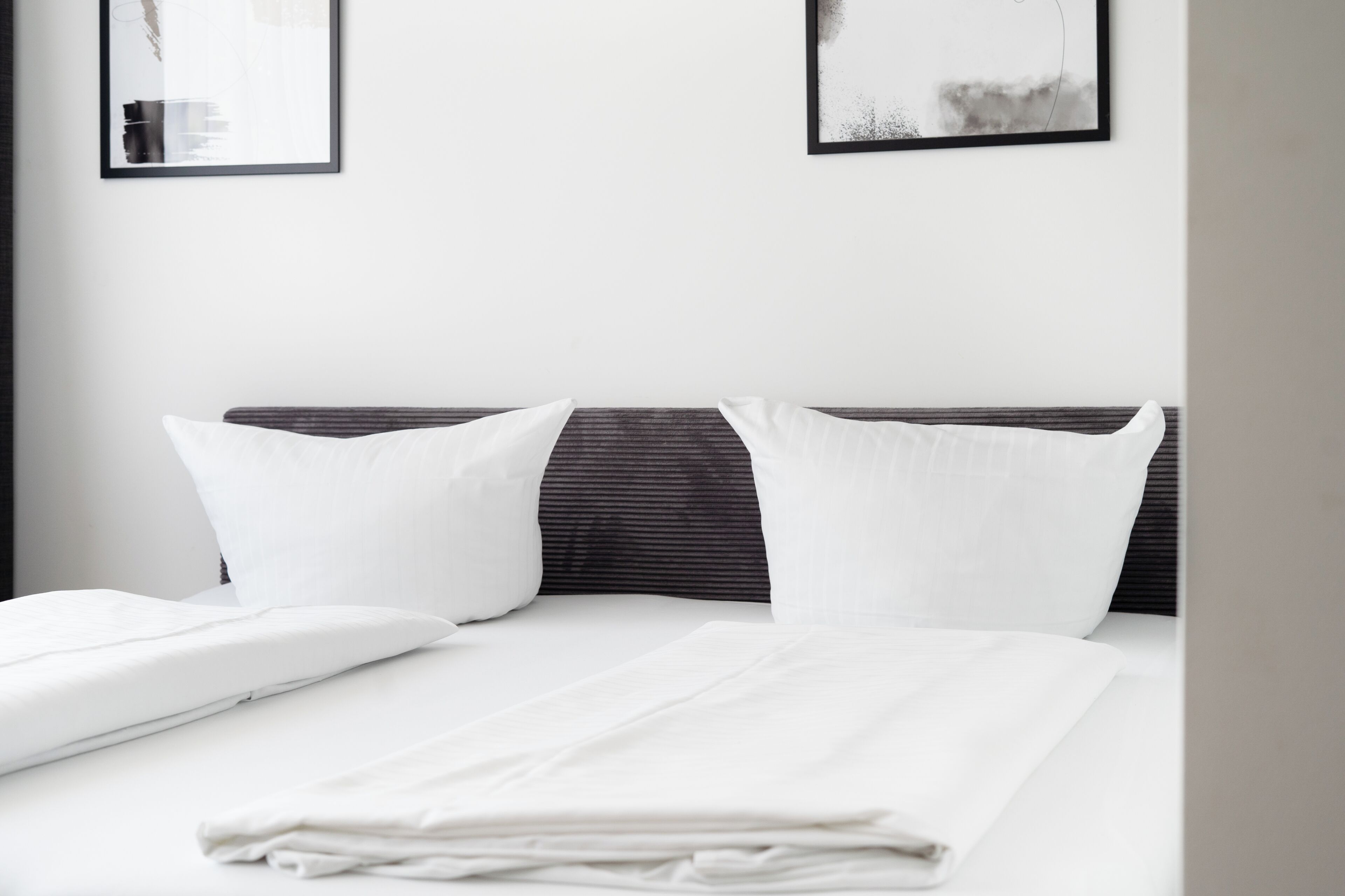 Una cama bien hecha con sábanas y almohadas blancas, cabecera oscura y dos cuadros en la pared.