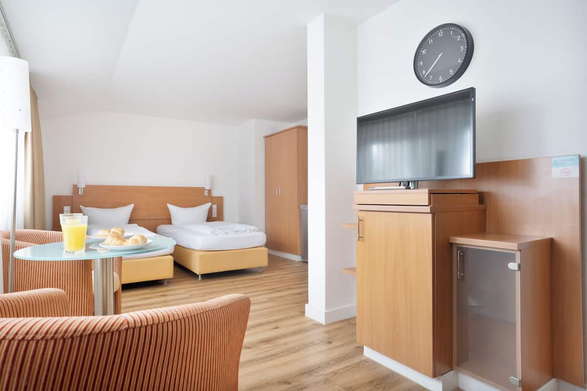 A hotel room with a bed, a TV, a small cabinet, a clock, and a table with a glass of juice and croissants.