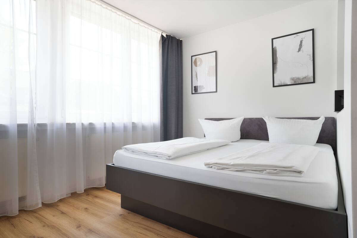 A modern hotel room with a large bed, a window with sheer curtains, and two framed artworks on the wall.