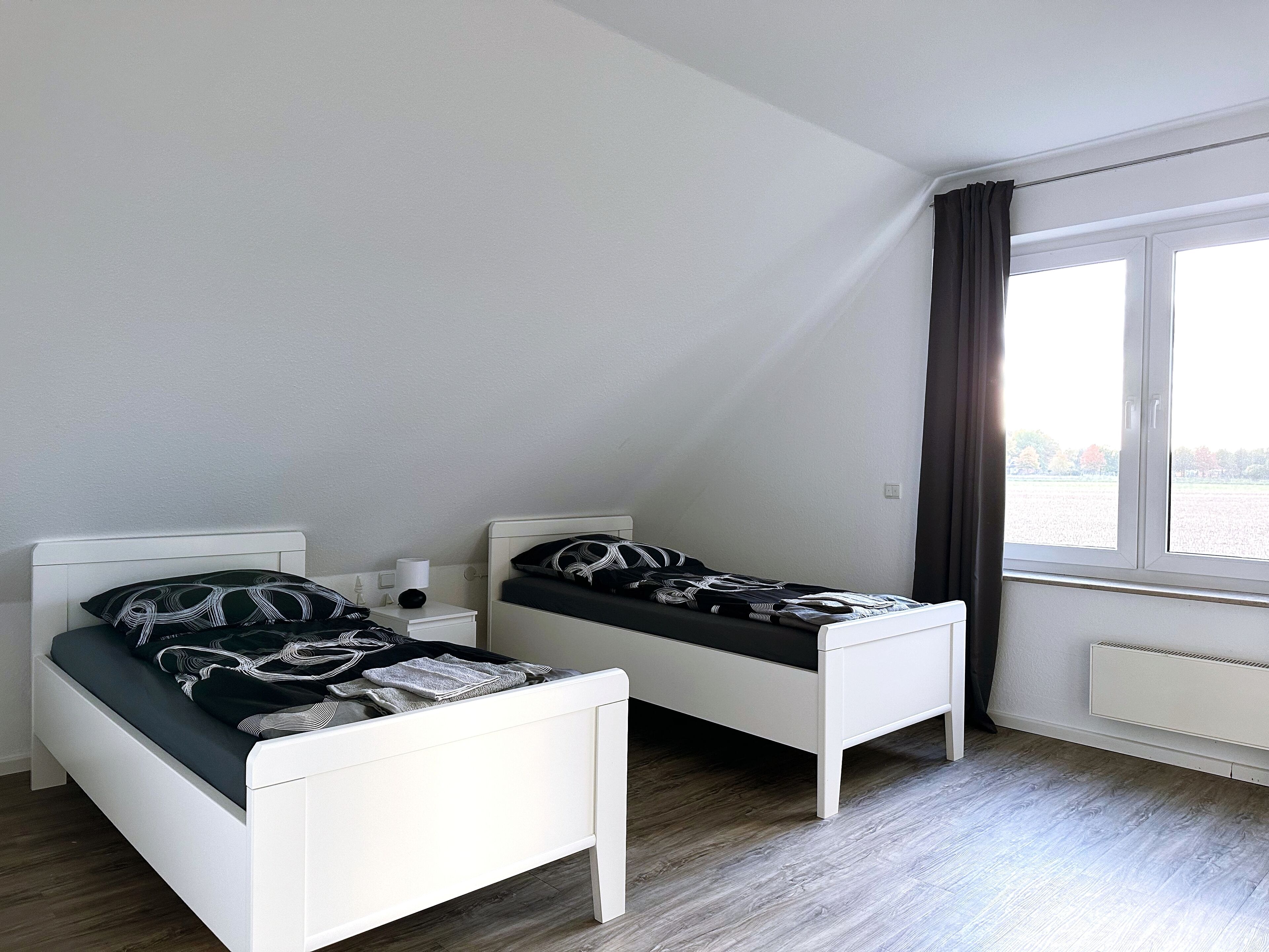 A small, clean room with two white beds, a window with curtains, and a radiator.