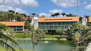 A building with a red roof and large windows, surrounded by palm trees and a small pond. - Flat 306 Villa Hípica Gravata (Gravata)