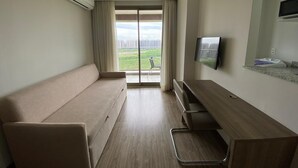 A hotel room with a sofa, a desk, a flat-screen TV, and a window with a view of buildings and greenery.