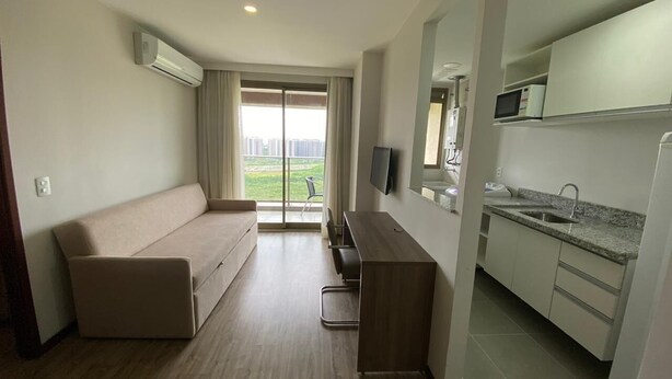 A compact hotel room with a sofa, a small desk, a flat-screen TV, a kitchen area with a microwave, and a sliding glass door leading to a balcony.