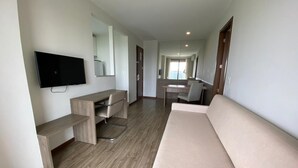 A modern hotel room with a sofa, a desk with a chair, a TV mounted on the wall, and a large mirror.