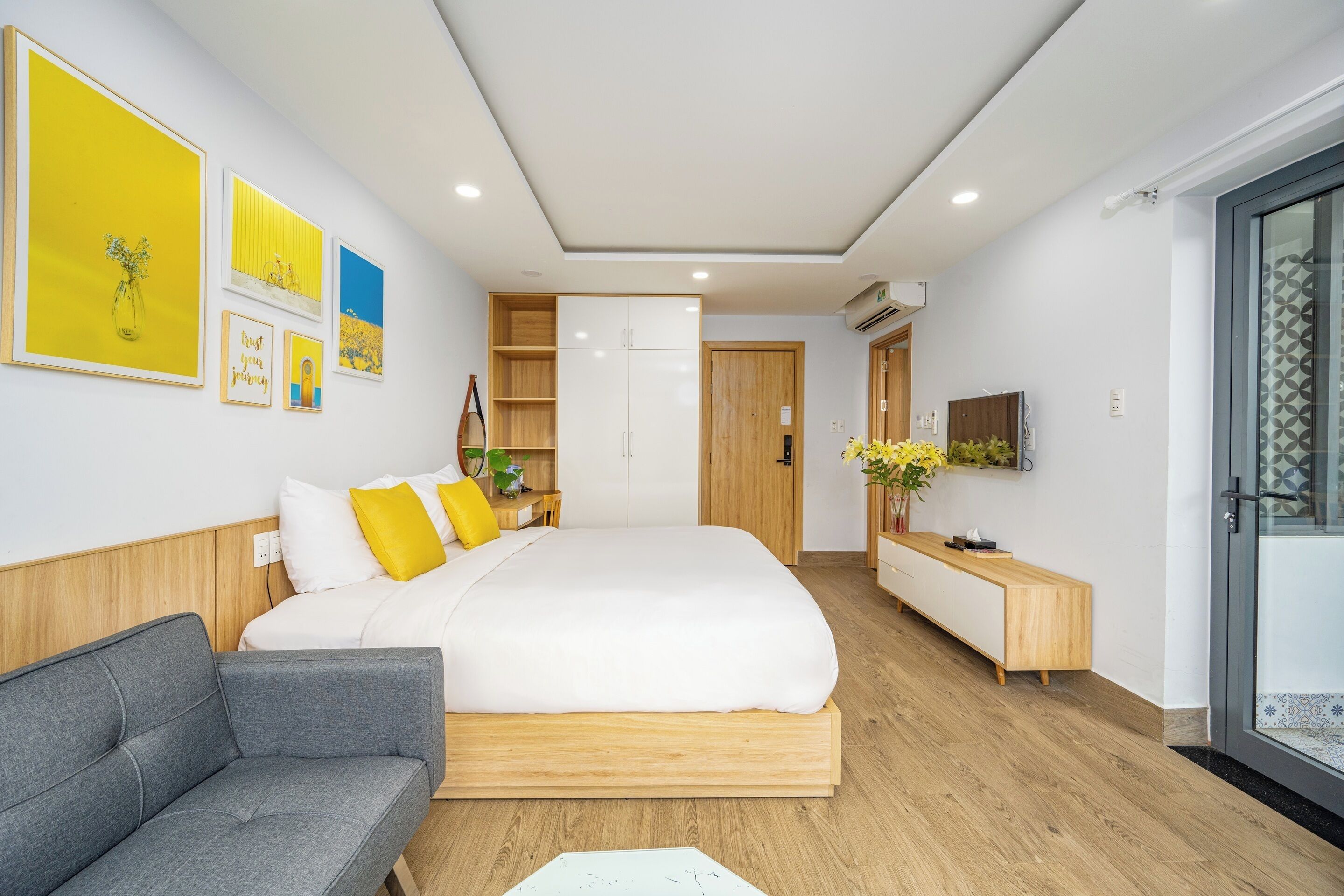 A modern bedroom with a large bed, a grey sofa, a wooden dresser, and a TV on a stand.