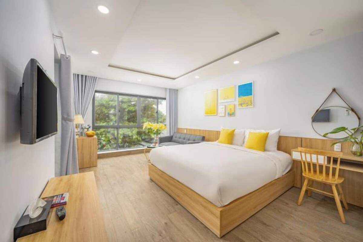 A modern hotel room with a large bed, a desk, a TV, and a window with a view of greenery.
