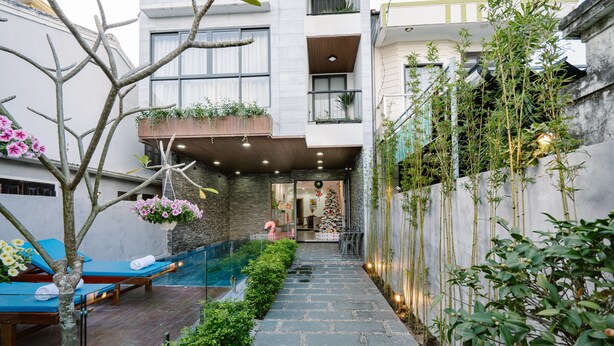 A modern building with a stone pathway leading to a pool area, surrounded by greenery and a bamboo fence. - LaSanta Villa (Da Nang)